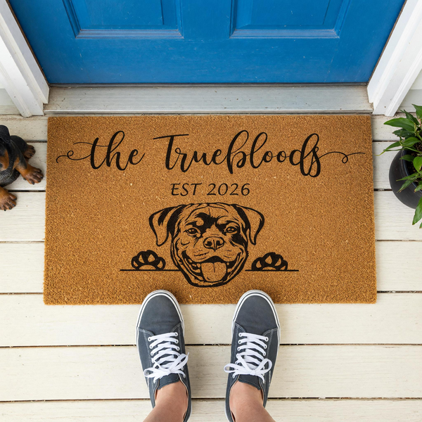 Personalized Rottweiler family doormat featuring a cute dog illustration and custom text on a natural coir mat, displayed on a white porch in front of a blue door with a person wearing gray sneakers standing on the mat for a