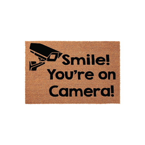 Smile You're on Camera doormat featuring a black security camera icon printed on a natural tan coir fiber mat, providing a humorous and functional way to greet guests while adding a touch of home security style to your front