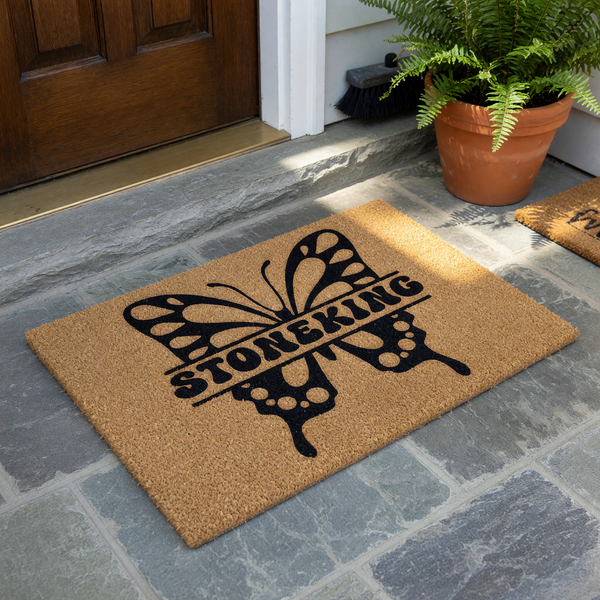 Personalized butterfly coir doormat featuring a large black butterfly silhouette with a split center for a custom family name, set against a natural brown textured fiber background for a welcoming front porch entrance decor piece.