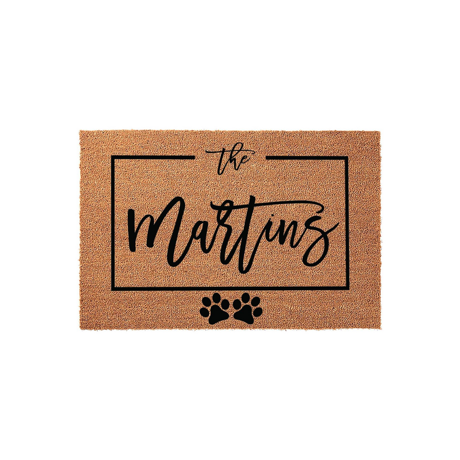 Personalized family name coir doormat featuring a rectangular black frame, elegant script text, and two cute paw prints at the bottom, creating a welcoming entrance for any pet-loving home on a clean, solid white studio
