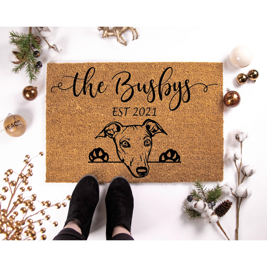Personalized Greyhound dog doormat featuring a cute illustration of a dog peeking over the edge, surrounded by festive Christmas decor like pine branches, cotton stems, and gold ornaments on a clean white background for a holiday