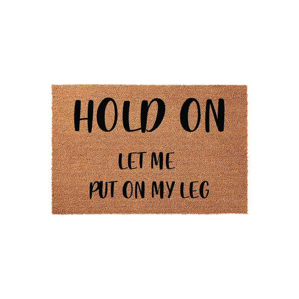 Humorous Hold On Let Me Put On My Leg doormat made of natural coir fiber with a plain white background, featuring bold black lettering that adds a funny and welcoming touch to any front porch or entryway decor for guests to enjoy.