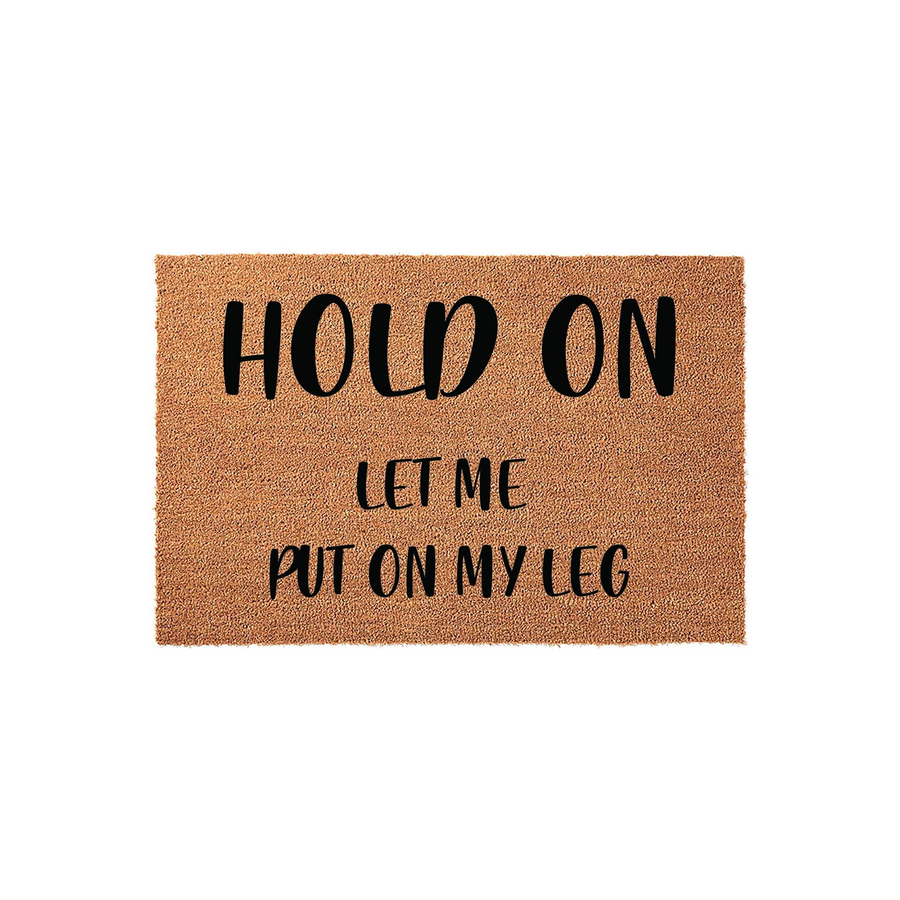 Humorous Hold On Let Me Put On My Leg doormat made of natural coir fiber with a plain white background, featuring bold black lettering that adds a funny and welcoming touch to any front porch or entryway decor for guests to enjoy.