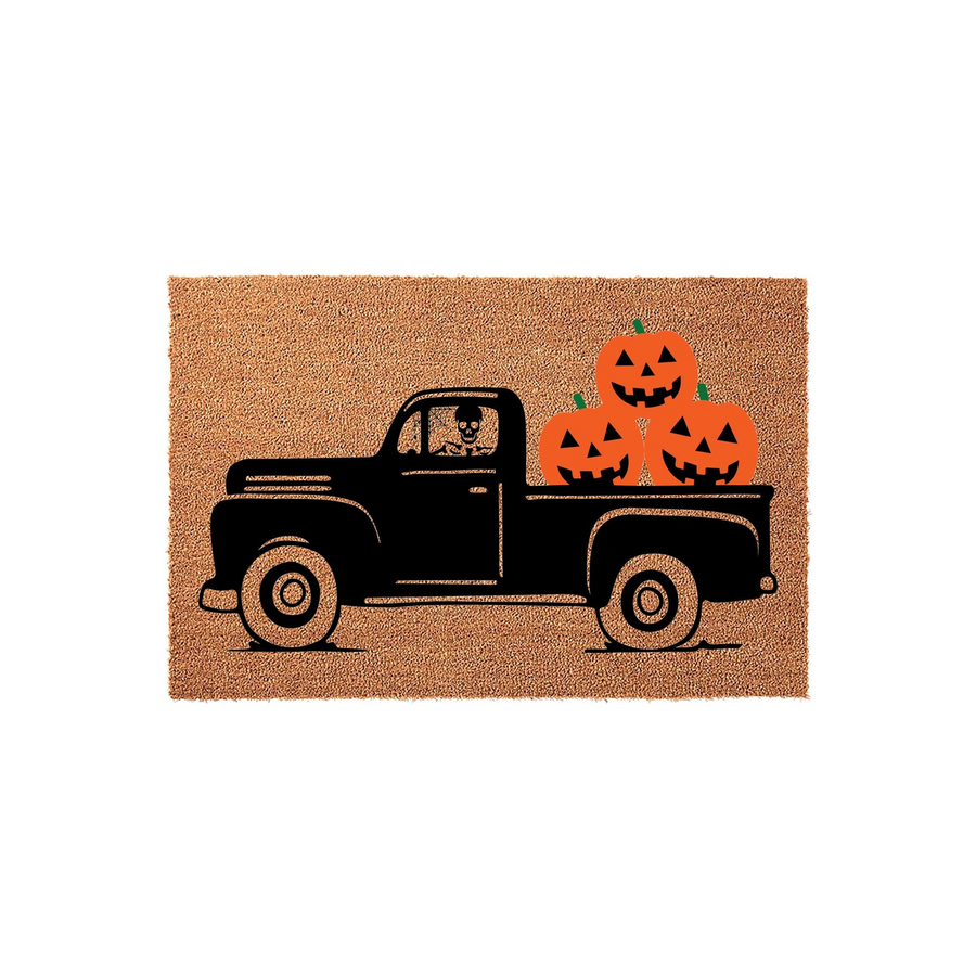 Halloween truck doormat featuring a black vintage pickup truck carrying three orange jack-o-lantern pumpkins in the bed, with a skeleton driver visible in the cab, all printed on a natural tan coir fiber outdoor welcome mat