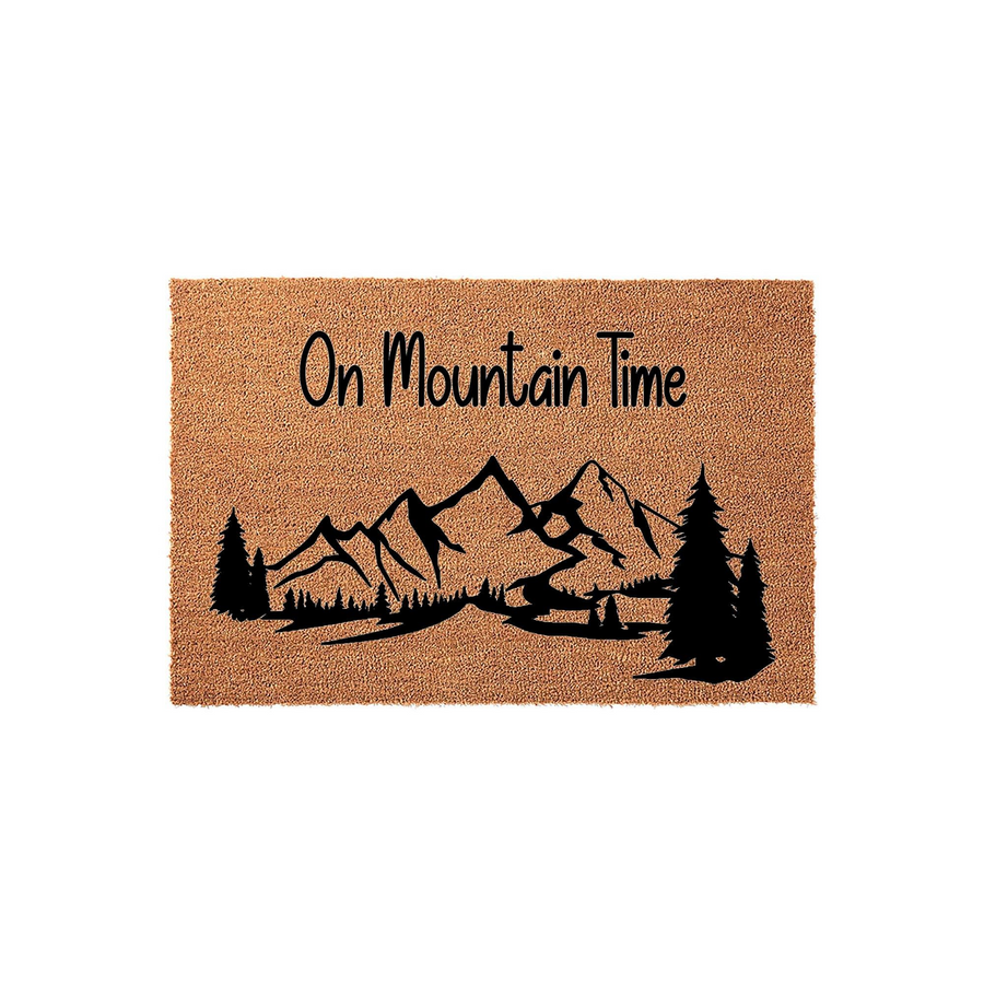 On Mountain Time coir doormat featuring a black printed graphic of a rugged mountain range and pine trees, set against a natural tan fiber background to provide a rustic outdoor aesthetic for your front porch or entryway decor.