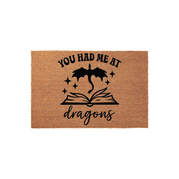 You Had Me At Dragons coir doormat featuring a black graphic of a flying dragon emerging from an open book with sparkling stars, set against a natural brown textured fiber background for a fantasy-themed home entryway decor piece.