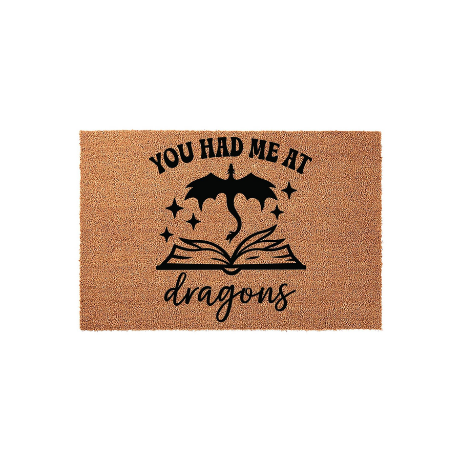 You Had Me At Dragons coir doormat featuring a black graphic of a flying dragon emerging from an open book with sparkling stars, set against a natural brown textured fiber background for a fantasy-themed home entryway decor piece.