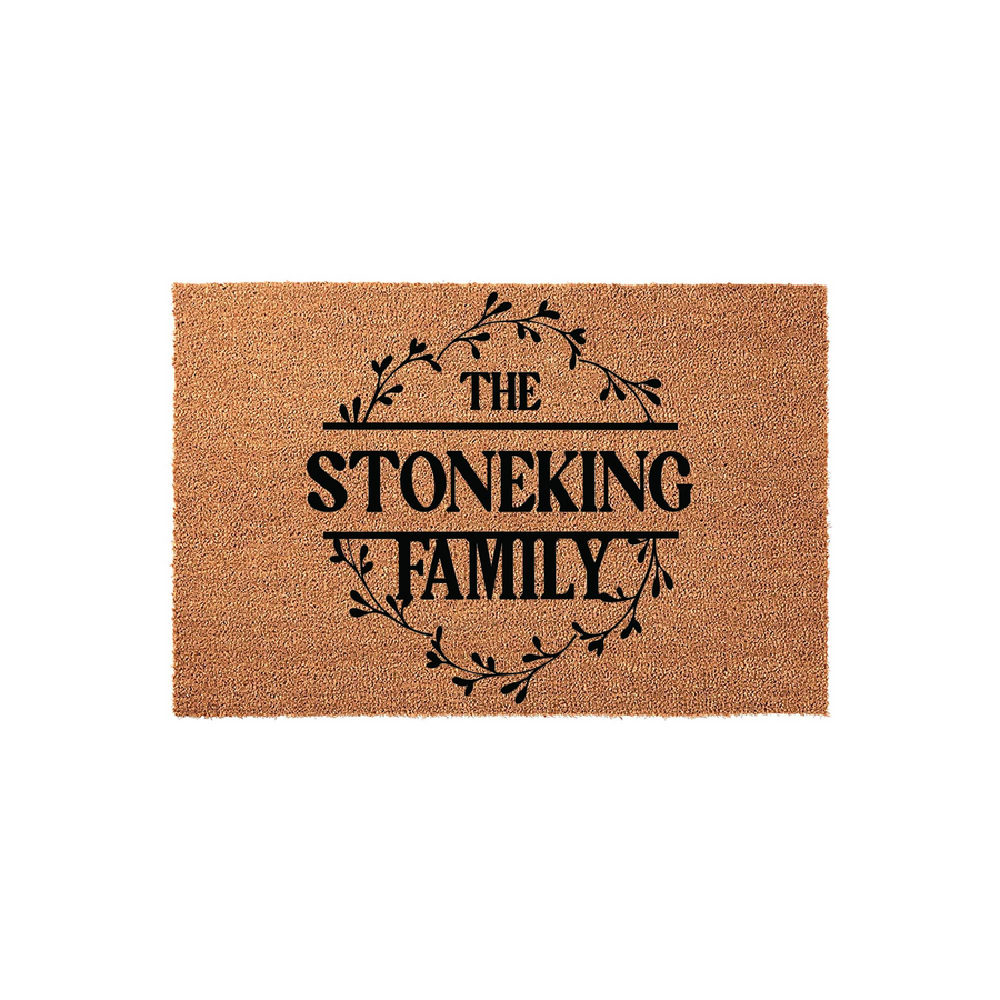 Personalized family name coir doormat featuring a rustic black leaf wreath design centered on a natural tan coconut fiber mat, providing a welcoming entrance decor piece for any home with custom last name text printed in bold