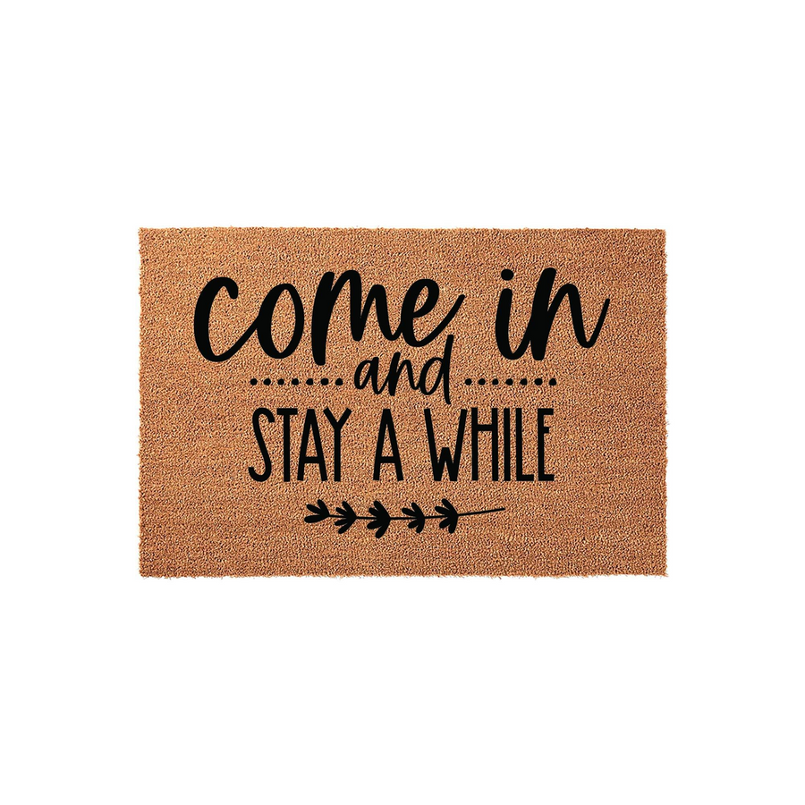 Come In and Stay A While coir doormat featuring a natural brown fiber texture and bold black typography, accented by a simple decorative leaf branch design at the bottom to create a welcoming entrance for your home front porch