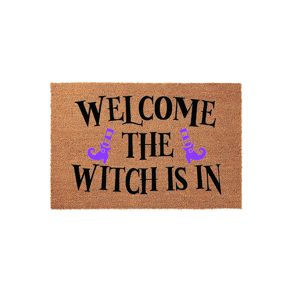 Welcome The Witch Is In Halloween Doormat featuring a natural coir texture with bold black lettering and two purple witch boots, creating a festive and spooky entryway decor piece for the fall season on a plain white background.