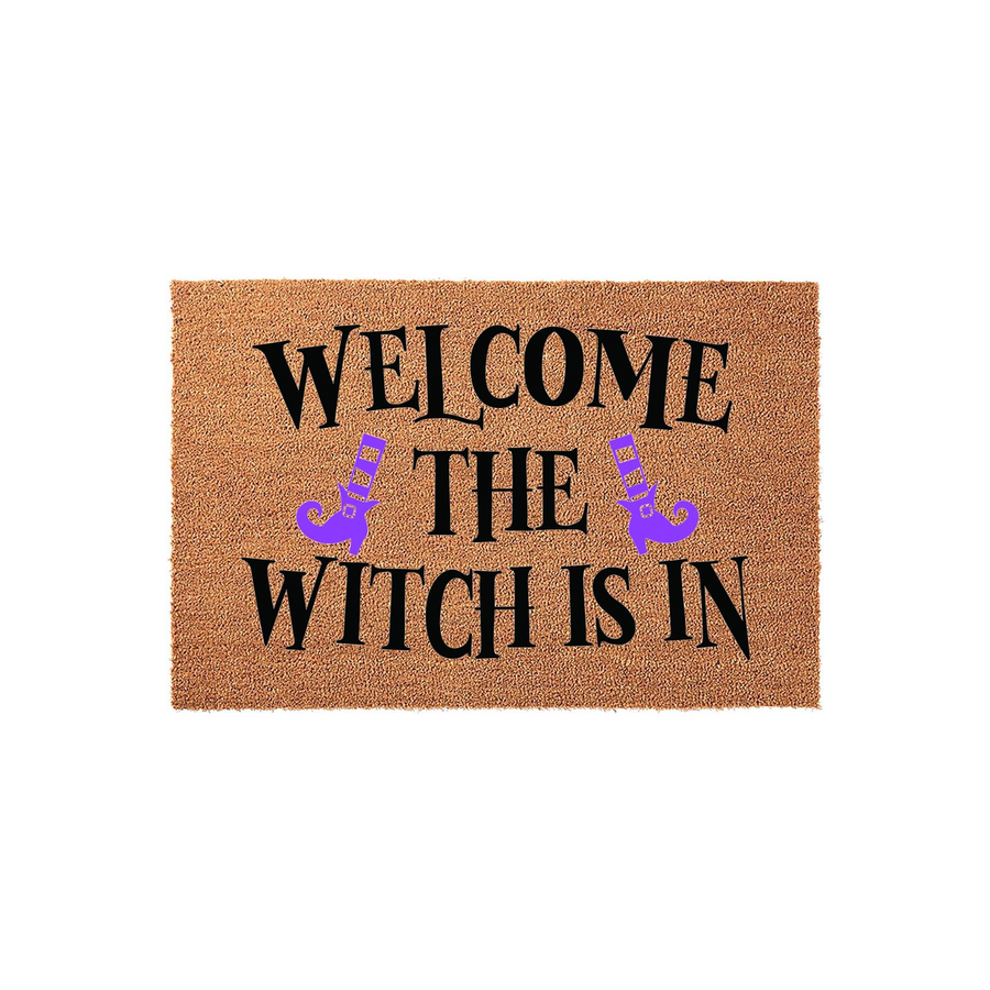 Welcome The Witch Is In Halloween Doormat featuring a natural coir texture with bold black lettering and two purple witch boots, creating a festive and spooky entryway decor piece for the fall season on a plain white background.