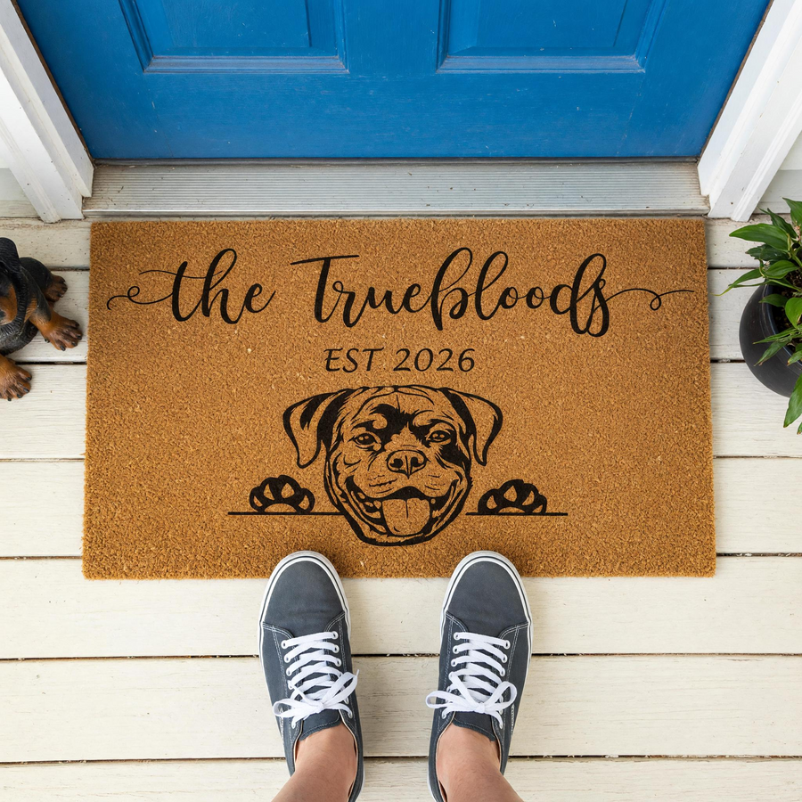 Personalized Rottweiler family doormat featuring a cute dog illustration and custom text on a natural coir mat, displayed on a white porch in front of a blue door with a person wearing gray sneakers standing on the mat for a