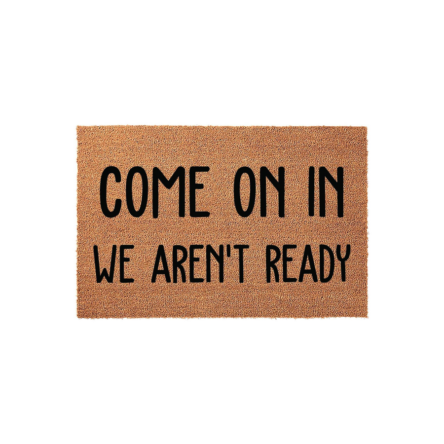 Funny Come On In We Aren't Ready coir doormat featuring bold black text printed on a natural tan fiber background, providing a humorous and welcoming touch to any front porch entryway or home decor setting for guests to enjoy.
