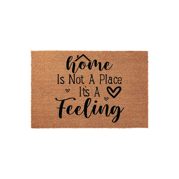 Home Is Not A Place It's A Feeling coir doormat featuring a natural tan fiber texture and bold black typography, designed as a welcoming front porch entrance rug with a charming house icon and small heart accents for a cozy home