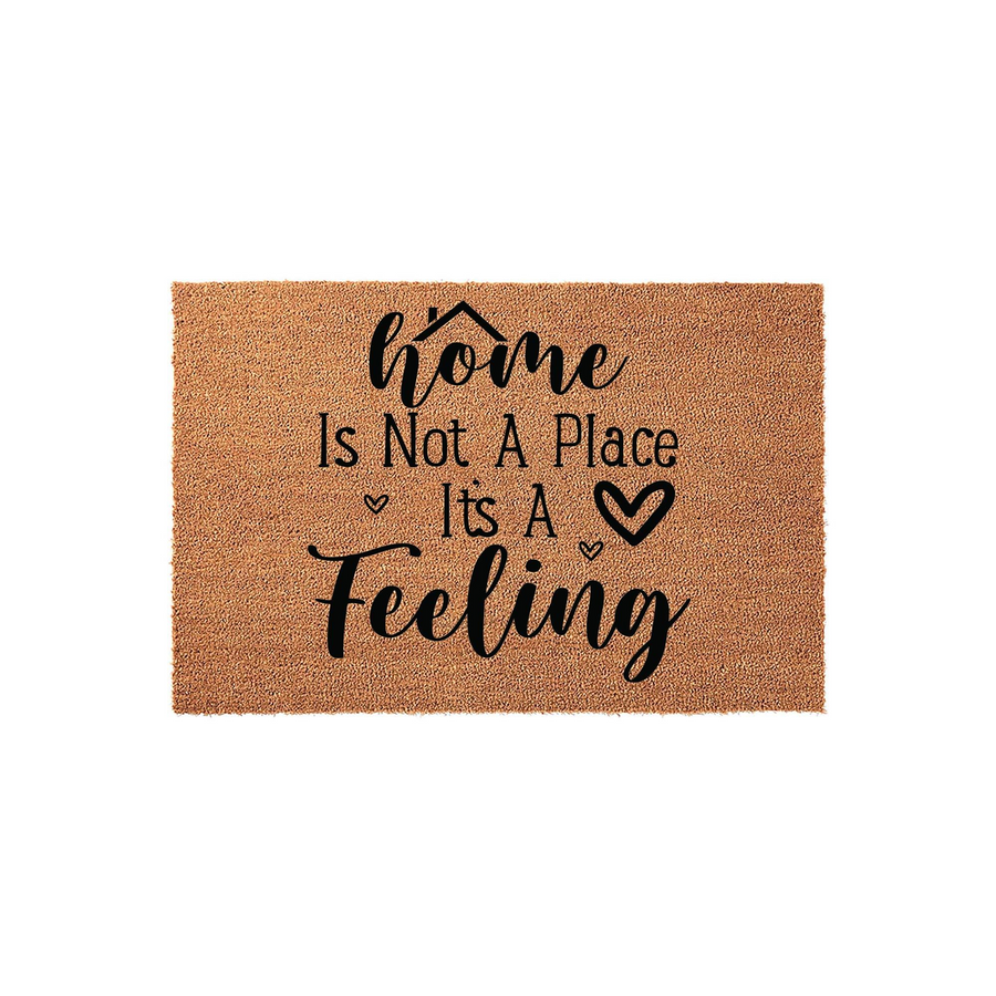 Home Is Not A Place It's A Feeling coir doormat featuring a natural tan fiber texture and bold black typography, designed as a welcoming front porch entrance rug with a charming house icon and small heart accents for a cozy home