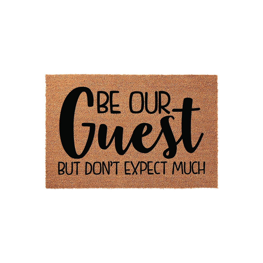 Funny Be Our Guest Doormat featuring a natural coir fiber texture with bold black text that reads Be Our Guest But Don't Expect Much, making it a humorous and welcoming addition to any front porch or entryway decor for your home.