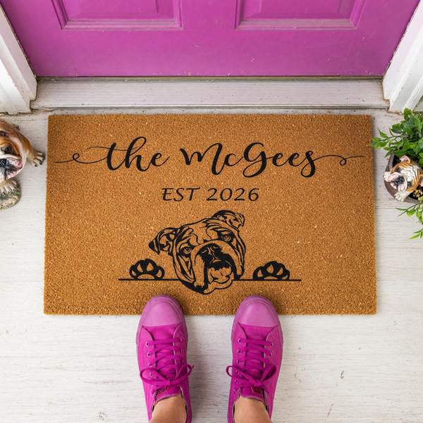 Personalized Bulldog Coir Doormat featuring a custom family name and established year, displayed on a porch in front of a bright purple door with a person wearing pink sneakers standing on the mat and decorative bulldog figurines