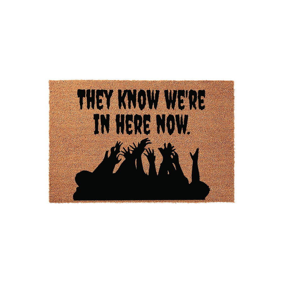 Funny Halloween doormat featuring a natural coir texture with black text that reads They Know We Are In Here Now, accompanied by a spooky silhouette of reaching zombie hands at the bottom for a festive and scary front porch
