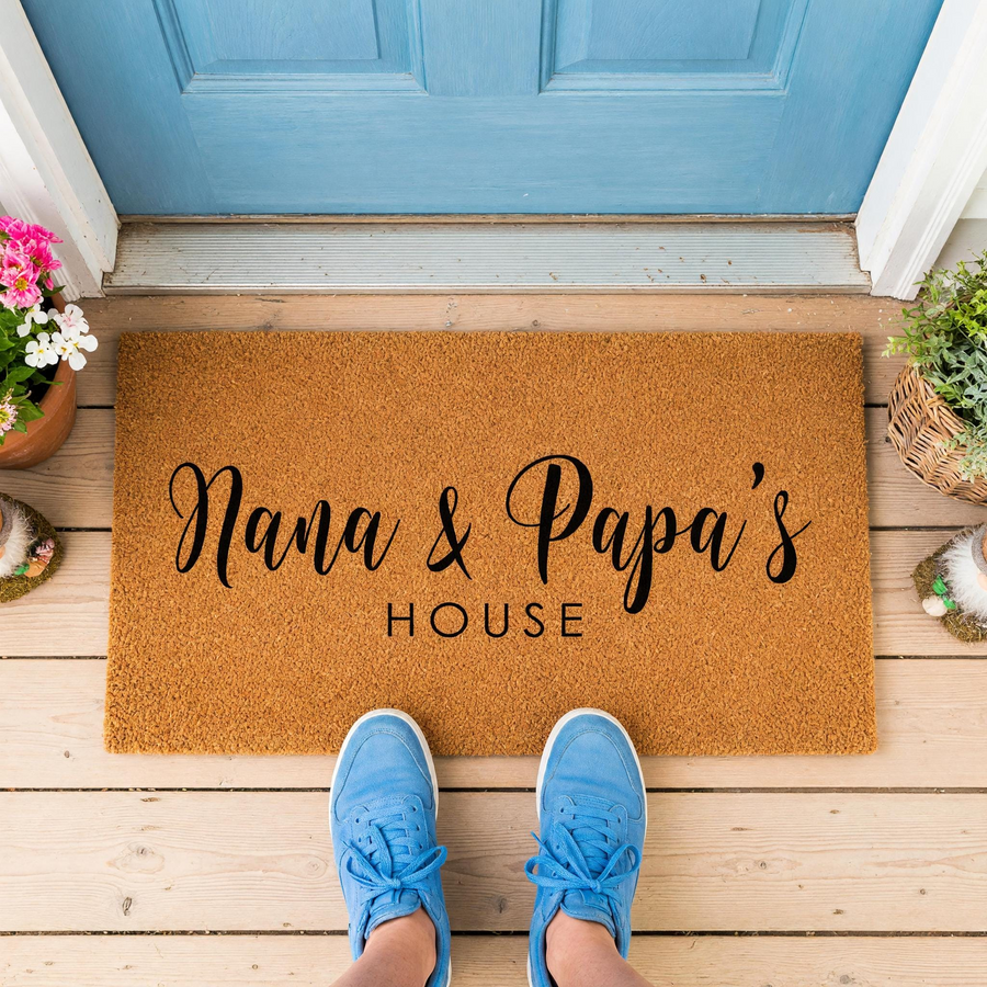 Nana and Papa's House personalized coir doormat featuring elegant black script lettering, displayed on a wooden porch in front of a blue door with a person wearing blue sneakers standing on the mat to welcome guests to the home.