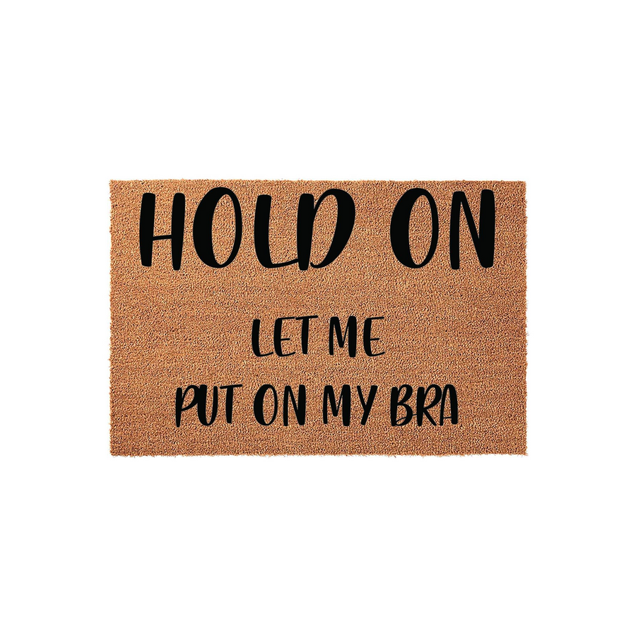Funny Hold On Let Me Put On My Bra coir doormat featuring bold black typography on a natural tan fiber background, making it a humorous and welcoming entrance mat for any home entryway or front porch decor setting.