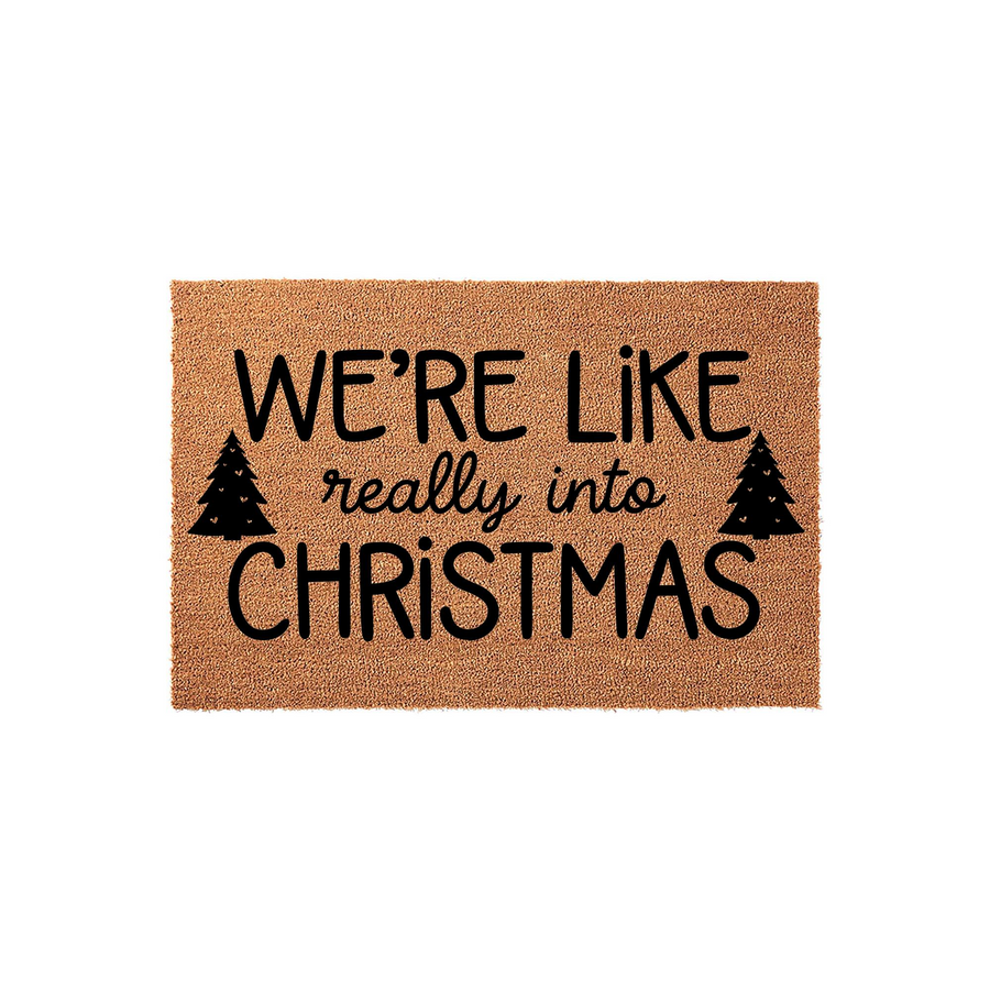 Christmas coir doormat featuring the text We are like really into Christmas in bold black lettering, accented by two small pine tree silhouettes on a natural brown fiber background for a festive holiday home entryway decor.