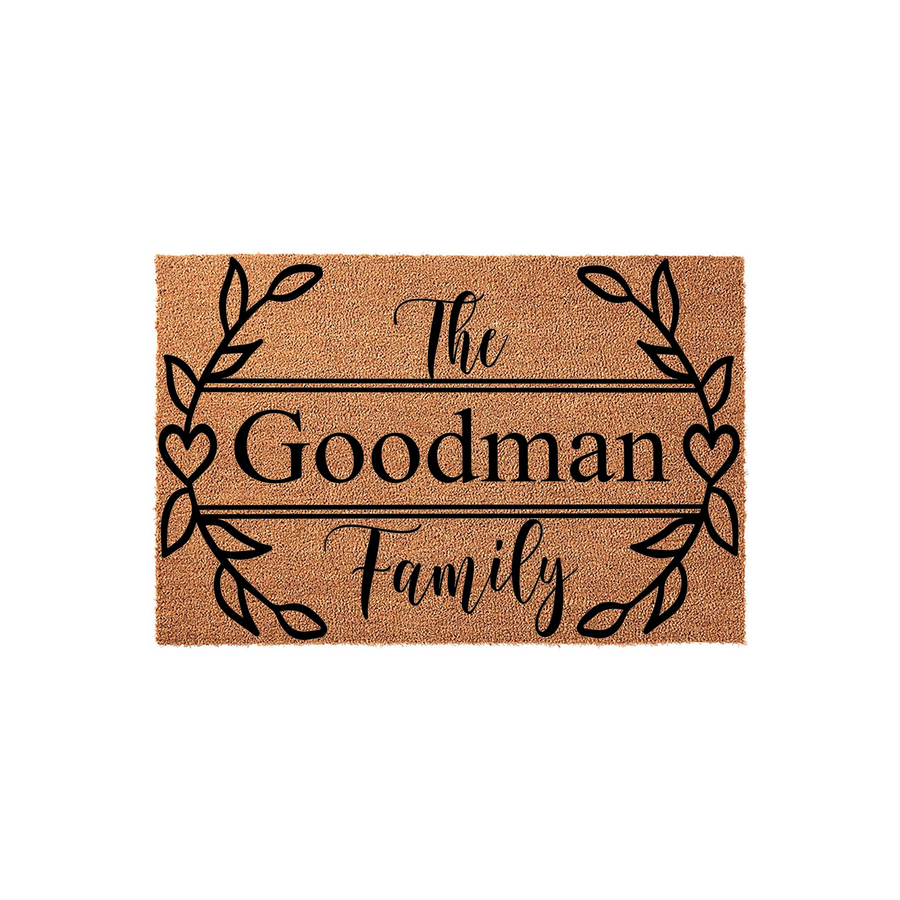 Personalized family name coir doormat featuring a decorative black leaf and heart wreath design printed on a natural tan fiber background, creating a welcoming entrance piece for any home or housewarming celebration.