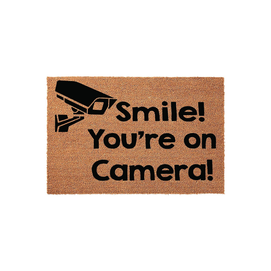 Smile You're on Camera doormat featuring a black security camera icon printed on a natural tan coir fiber mat, providing a humorous and functional way to greet guests while adding a touch of home security style to your front