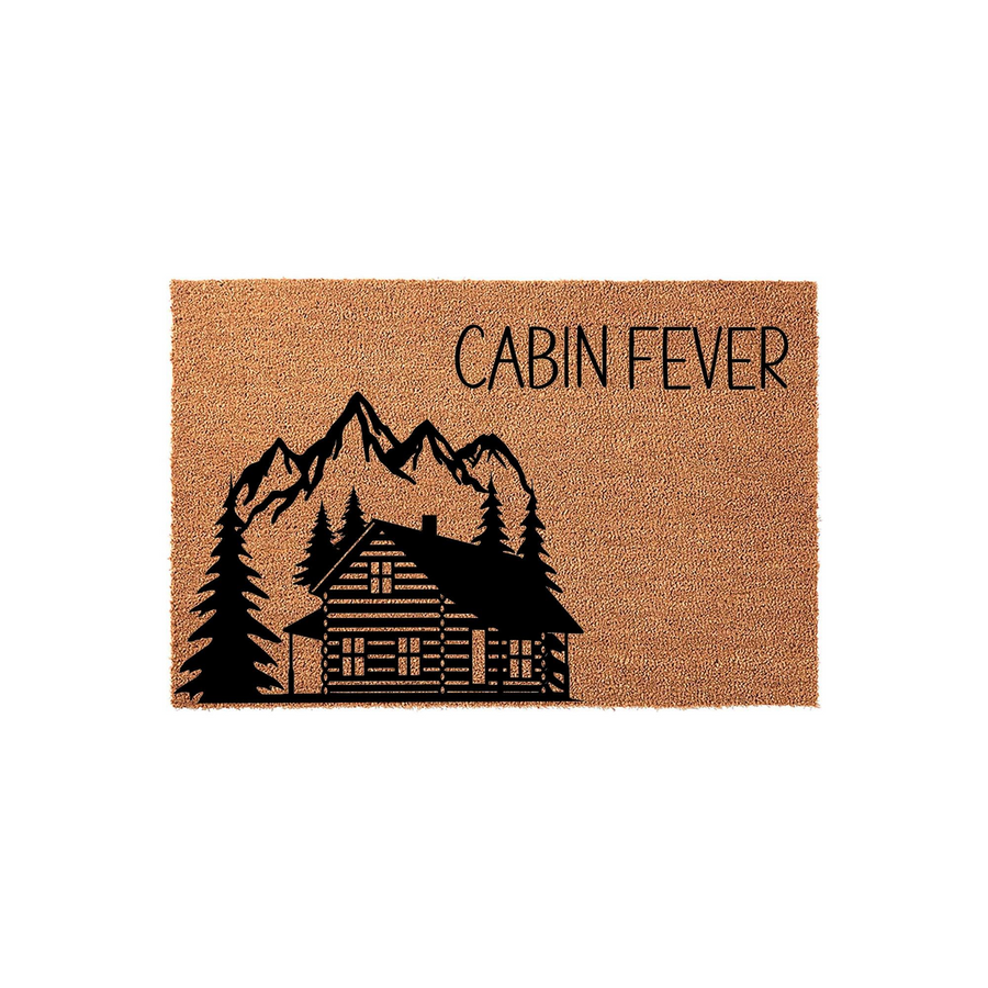 Cabin Fever natural coir doormat featuring a black printed graphic of a rustic log cabin nestled in front of tall pine trees and jagged mountain peaks, set against a clean white background for a cozy outdoor entryway decor