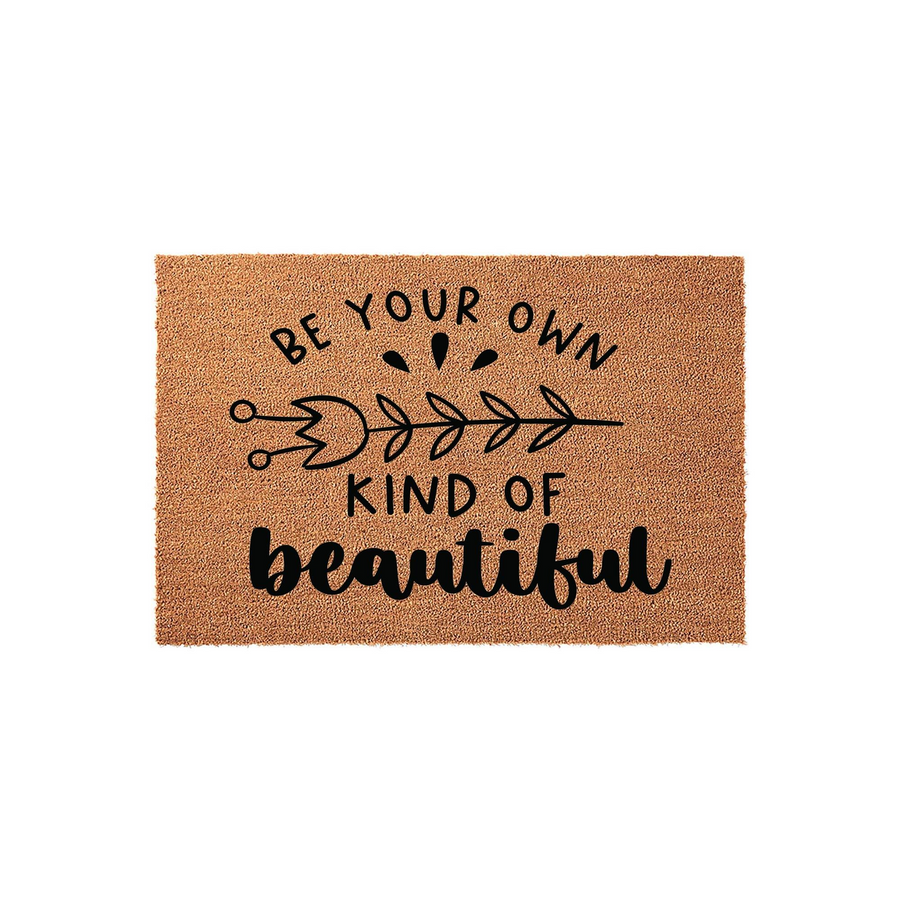 Be Your Own Kind of Beautiful Coir Doormat featuring a natural brown fiber texture with black printed text and a decorative leaf branch design, centered on a plain white background for a clean and welcoming entryway decor