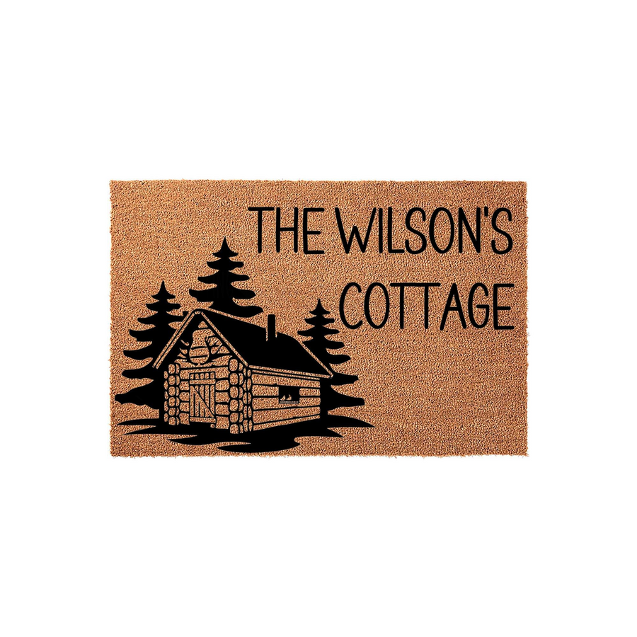 Personalized cabin doormat featuring a rustic log cabin and pine tree illustration on a natural coir background, customized with a family name and the word cottage in bold black lettering for a welcoming outdoor entryway decor