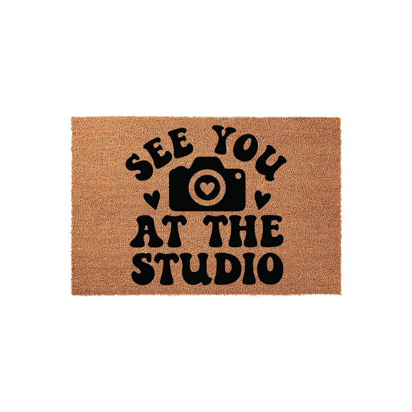 See You At The Studio photography doormat featuring a natural coir texture with bold black lettering and a camera icon with a heart lens, creating a welcoming entrance for a professional photography studio or home office space.