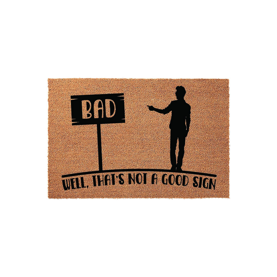 Funny Welcome Doormat featuring a silhouette of a man pointing at a sign that says BAD, with the text Well, That's Not A Good Sign printed below on a natural tan coir fiber background for a humorous entryway decor statement piece.