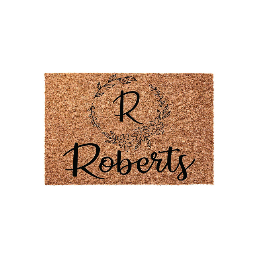 Personalized family name coir doormat featuring a decorative floral wreath design surrounding a large initial letter, providing a welcoming and rustic aesthetic for your front porch entrance on a natural tan fiber background.