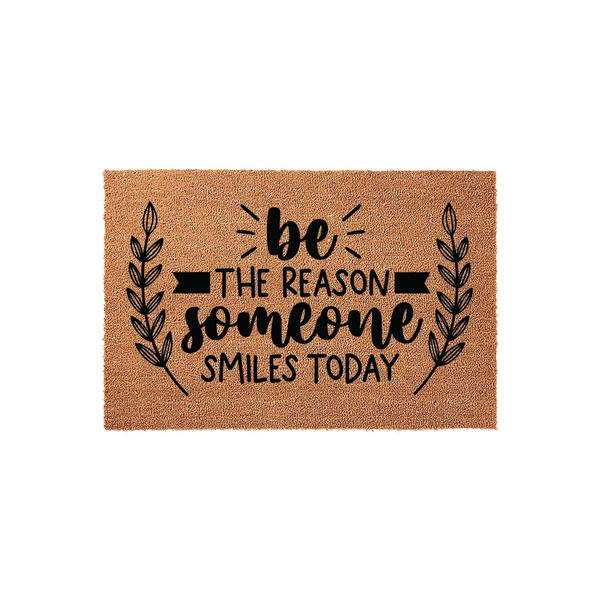Be The Reason Someone Smiles Today coir doormat featuring a natural brown fiber texture and black printed text with decorative leaf branches on the sides, creating a welcoming entryway decor piece for your front porch or home