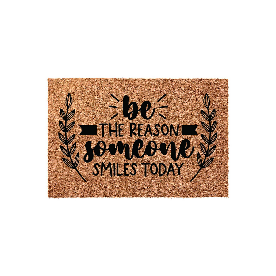 Be The Reason Someone Smiles Today coir doormat featuring a natural brown fiber texture and black printed text with decorative leaf branches on the sides, creating a welcoming entryway decor piece for your front porch or home