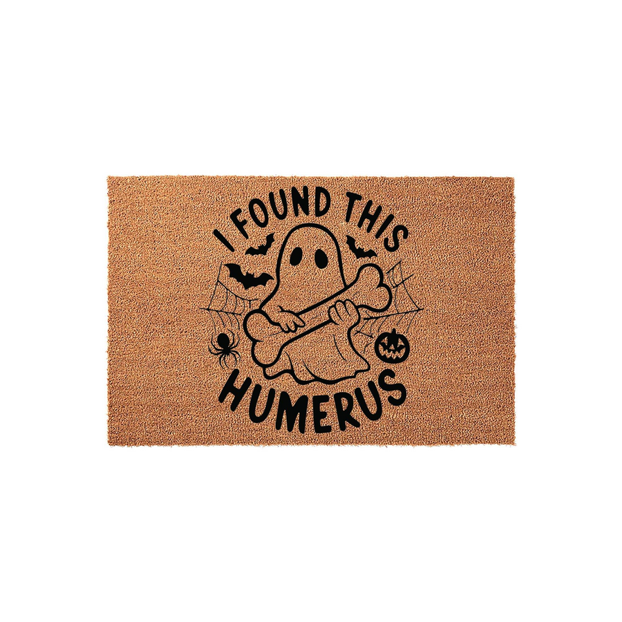 Funny Halloween doormat featuring a cute ghost holding a bone with the text I Found This Humerus, set against a natural coir fiber background with black spider, bat, and pumpkin illustrations for a festive seasonal entryway decor.