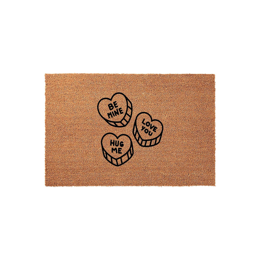 Valentine Conversation Heart Doormat featuring three candy heart illustrations with the phrases Be Mine, Love You, and Hug Me printed in black on a natural brown coir fiber mat, creating a festive entryway decor for the holiday