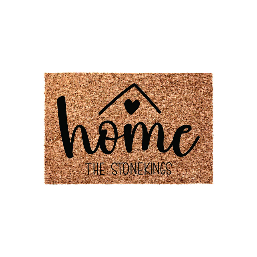 Personalized coir doormat featuring the word home in black script with a heart icon above the letter o, displayed on a plain white background with space for a custom family name to be printed underneath the main design.