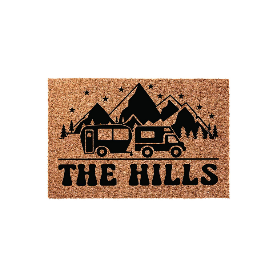 Personalized camping doormat featuring a black graphic of a truck pulling a travel trailer under mountain peaks and stars, with custom family name text printed on a natural tan coir fiber background for a rustic outdoor home
