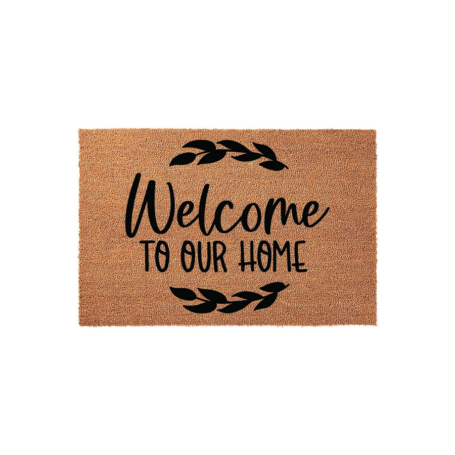 Welcome To Our Home coir doormat featuring a natural tan fiber texture and black printed text with decorative leaf accents, providing a rustic and inviting entryway decor piece for your front porch or house entrance.