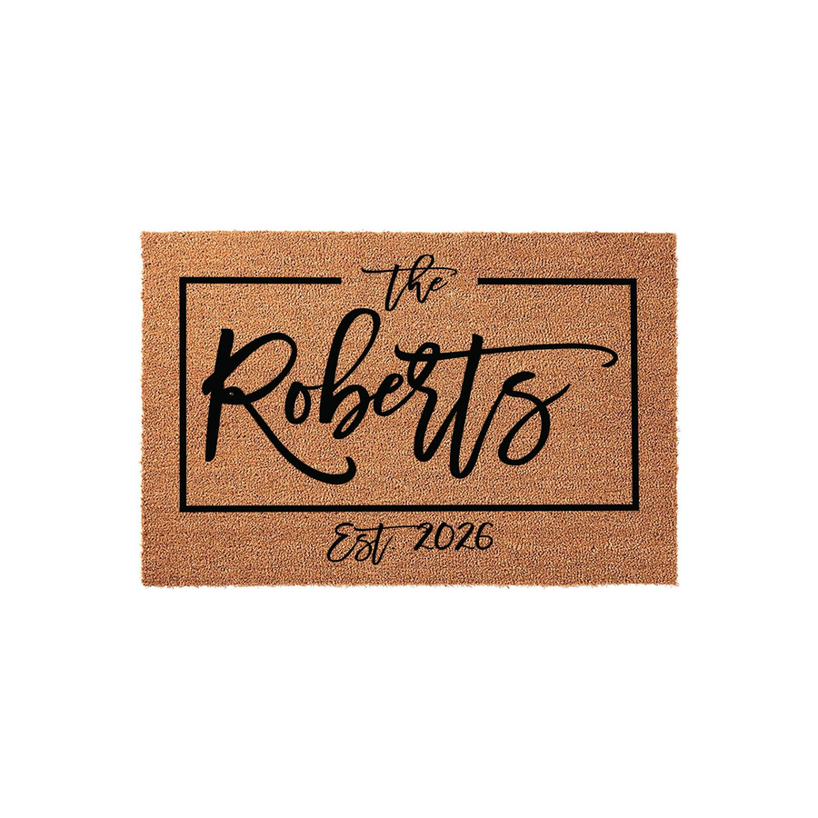 Personalized family name coir doormat featuring a rectangular black border with elegant script text that includes the family name and established year, set against a natural tan textured coconut fiber background for a front porch.