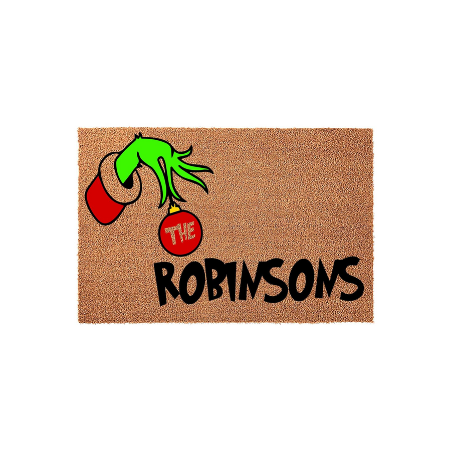 Personalized Christmas doormat featuring a green hand holding a red ornament with the word The and a custom family name printed in black, set against a natural brown coir texture for a festive holiday home entrance decoration.