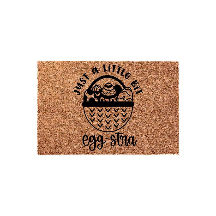Easter Egg-stra Coir Doormat featuring a natural brown fiber texture with a black printed design of a basket filled with patterned eggs and the playful text Just a Little Bit Egg-stra for a festive spring home entryway decoration.