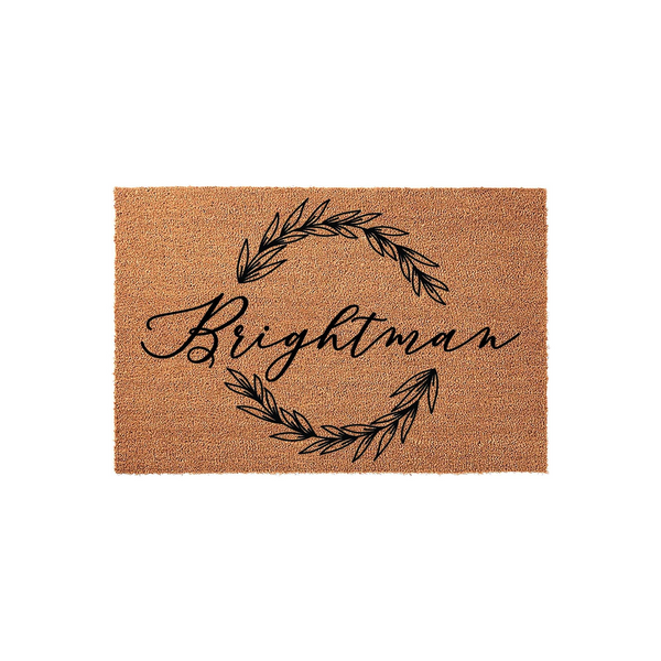 Personalized family name coir doormat featuring a minimalist black leaf wreath design centered on a natural brown fiber background, providing a welcoming and stylish entrance decor piece for any home front porch or entryway.