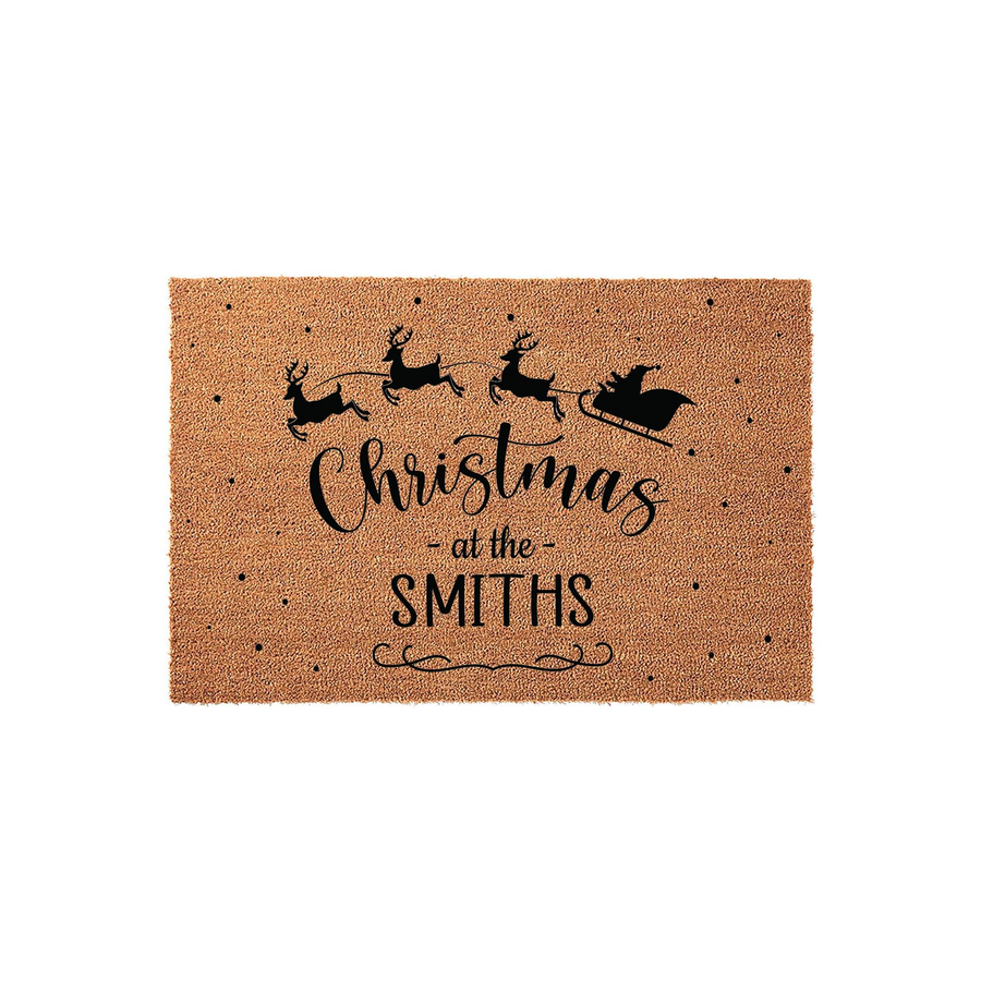 Personalized Christmas doormat featuring a black silhouette of Santa and his reindeer flying across a natural coir fiber mat, with festive text that reads Christmas at the Smiths and a decorative flourish on a plain white