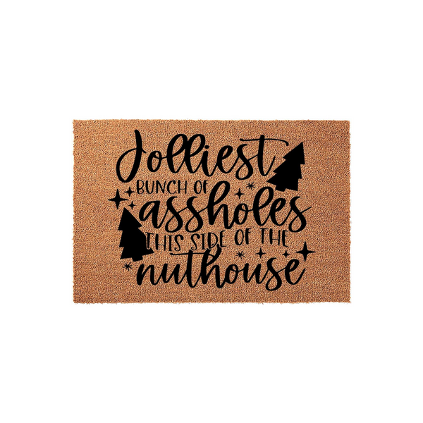 Funny Christmas doormat made of natural coir fiber, featuring black text that reads Jolliest Bunch of Assholes This Side of the Nuthouse, accented with decorative pine tree silhouettes and stars on a plain white studio background.