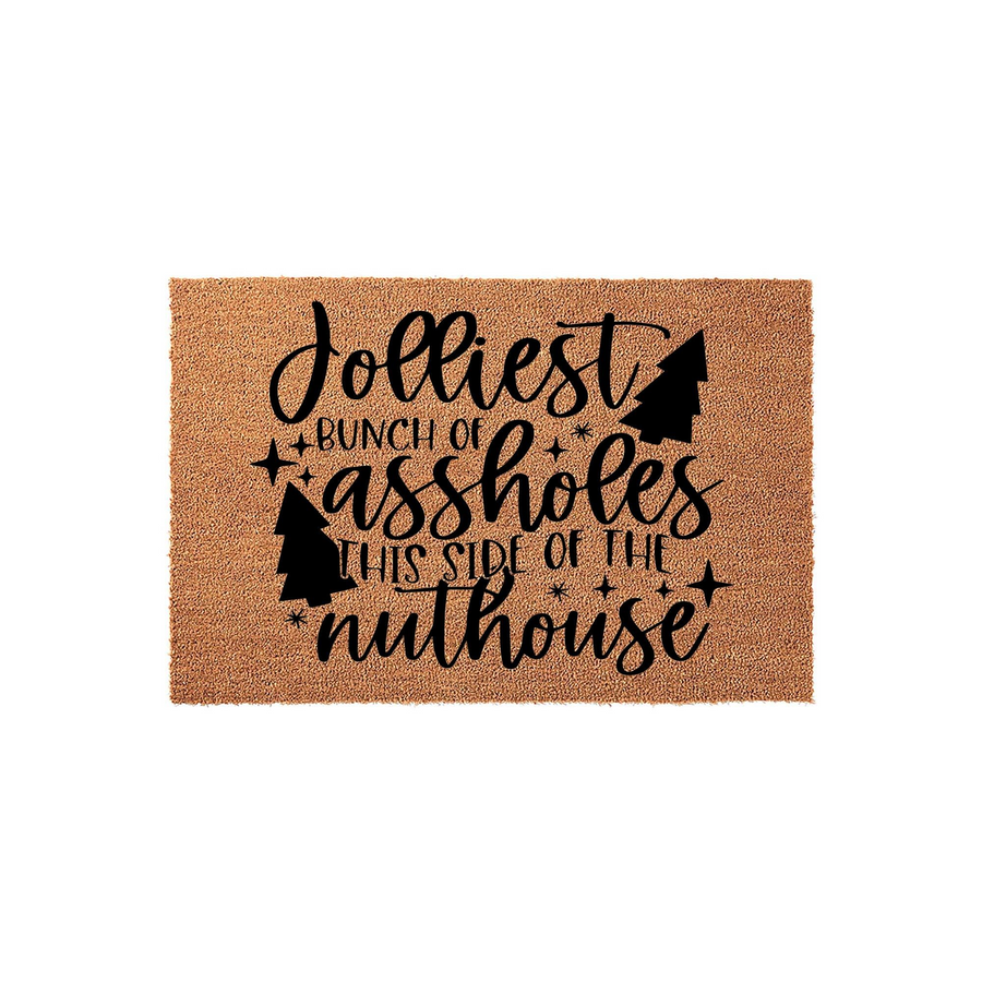 Funny Christmas doormat made of natural coir fiber, featuring black text that reads Jolliest Bunch of Assholes This Side of the Nuthouse, accented with decorative pine tree silhouettes and stars on a plain white studio background.