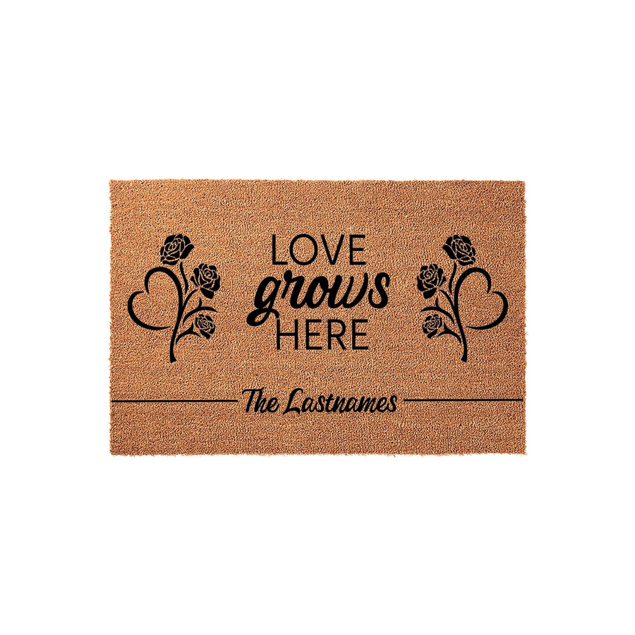 Personalized Love Grows Here coir doormat featuring black printed text and floral heart designs on a natural tan fiber background, providing a welcoming entrance for any family home with space for a custom last name at the bottom.