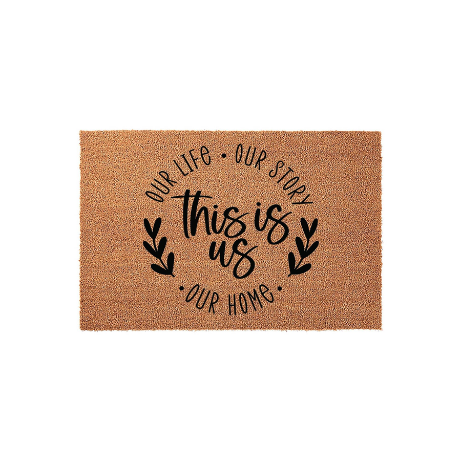 This Is Us Coir Doormat featuring a natural tan fiber texture with black printed text that reads Our Life, Our Story, Our Home, surrounding the central phrase This Is Us, accented by simple leaf illustrations on a clean white