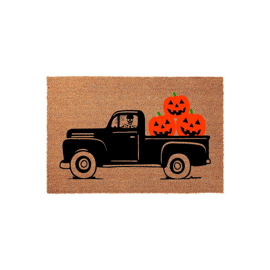 Halloween Truck Coir Doormat featuring a black vintage pickup truck carrying a stack of bright orange jack-o-lantern pumpkins, with a spooky skeleton driver visible in the cab against a natural tan fiber background for fall home